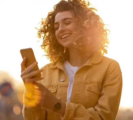 A woman with the sun behind her looking at her phone smiling.