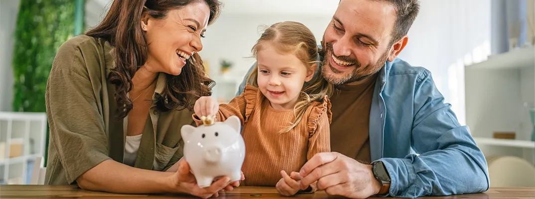 Two parents helping their daughter add coins to a piggy bank.