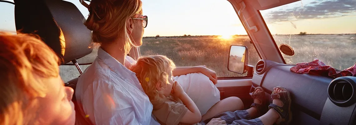 A woman sitting in the front seat of a car with two kids. All three are watching the sun set