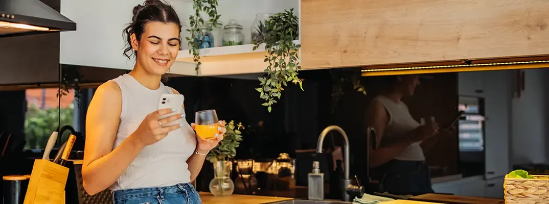 A woman looking at her phone holding a glass of orange juice in a kitchen