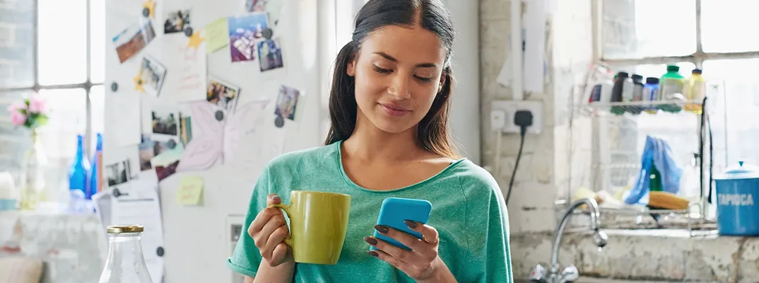 Woman with a coffee cup standing in a kitchen looking at her smartphone.