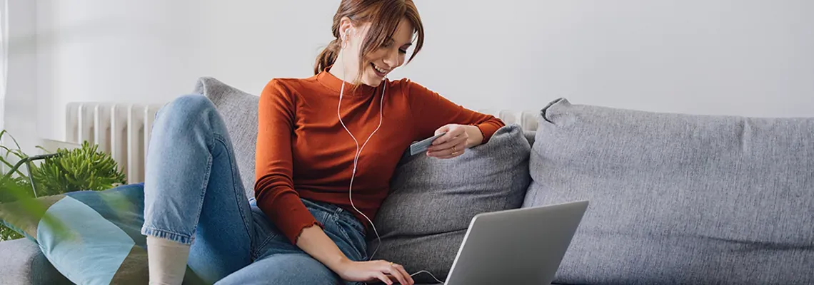 A woman with headphones looking at her credit card in front of a laptop