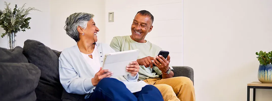 An elderly couple on a couch with their ipad and phone smiling at each other.