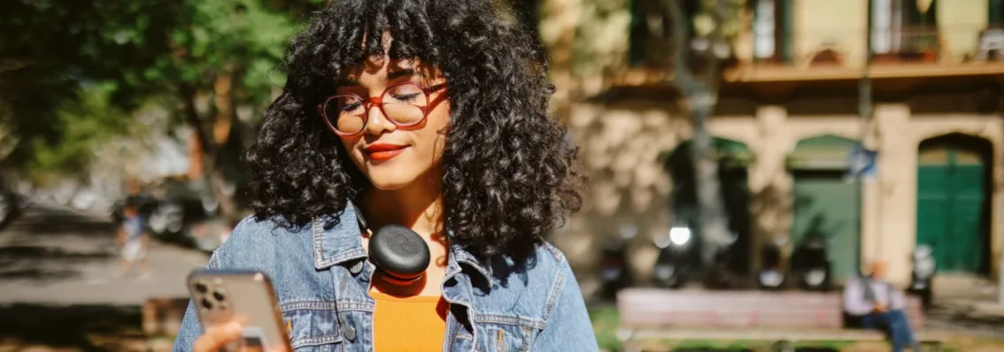 A woman standing in the sun, in a park, looking at her phone