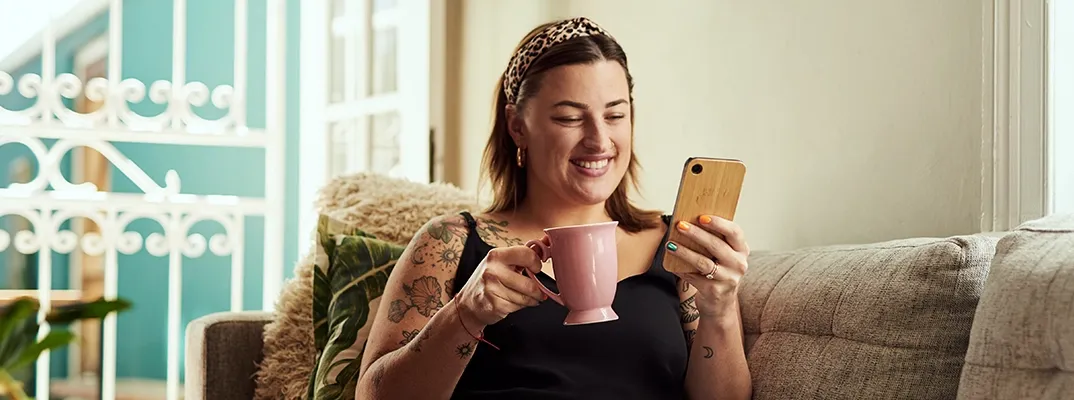 Woman with a coffee cup sitting on a couch looking at her smartphone.