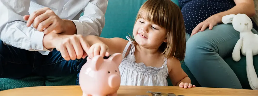 A small girl adding coins to a piggy bank.