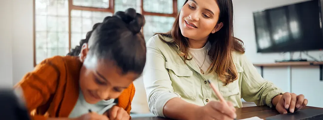 A girl is writing at a table while a woman next to her looks on.