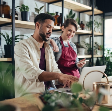 Man and woman looking at a laptop, standing in a room with potted plants on tables and shelves