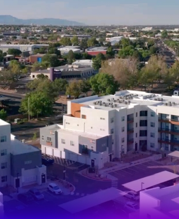 Drone view of a modern apartment community with multiple residential buildings, parking areas, and nearby commercial and residential properties.