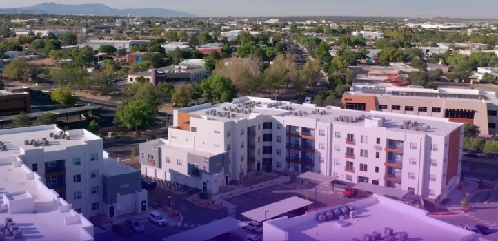 Drone view of a modern apartment community with multiple residential buildings, parking areas, and nearby commercial and residential properties.