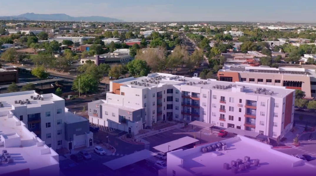 Drone view of a modern apartment community with multiple residential buildings, parking areas, and nearby commercial and residential properties.