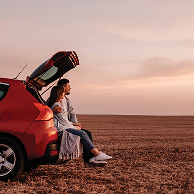 Young couple sitting on their car's open hatchback watching sunset in a field