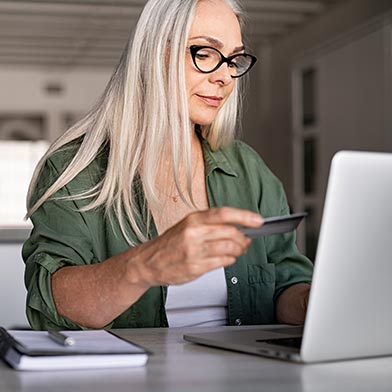 Older woman with glasses looking down at credit card while paying bills using her laptop