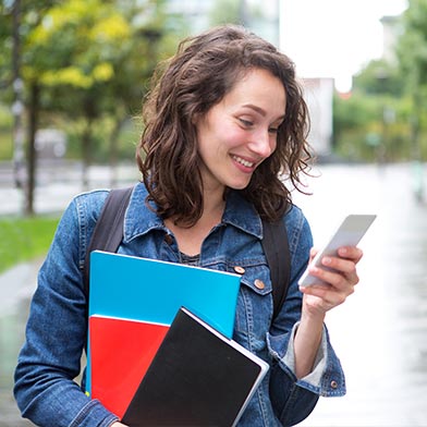 College girl with backpack carrying notebooks and looking down at cellphone