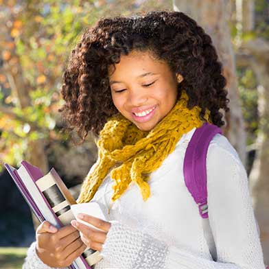 Young woman carrying school books and smiling at her smart phone.