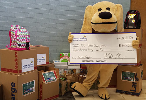 Sandy posing by boxes holding donated school items and holding a check for monetary donations.