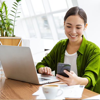 Smiling woman sitting at desk in front of laptop and looking at cell phone