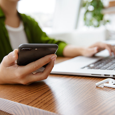 woman holding a cell phone and typing on a laptop