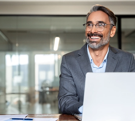 A well-dressed, smiling man sitting at a desk with a laptop