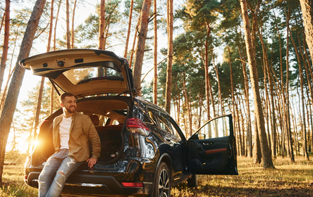 A man sitting in the open back hatch of a small SUV in a forest